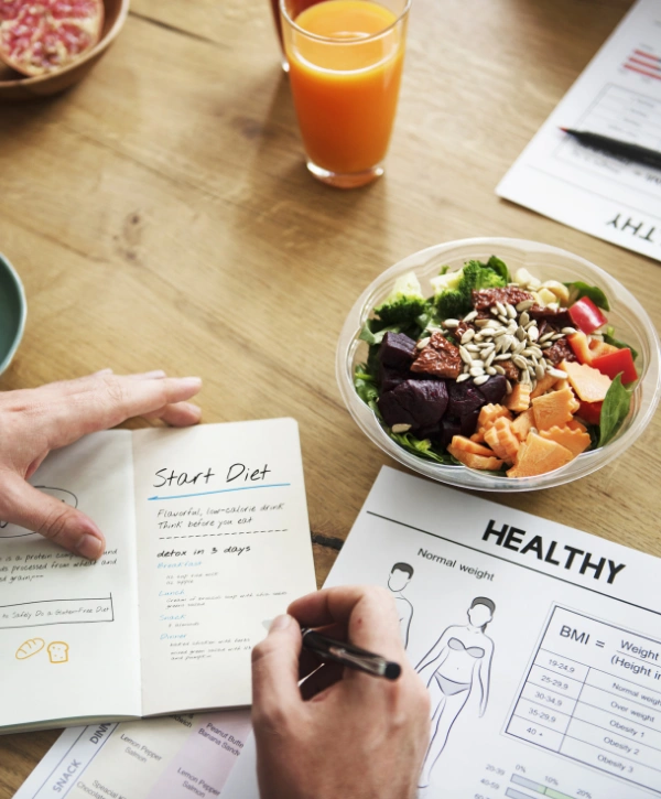 Person filling out daily diet beside a bowl of vegetables and grains, along with a glass of orange juice - Balancing Out Nutritional Plan for Better Recovery