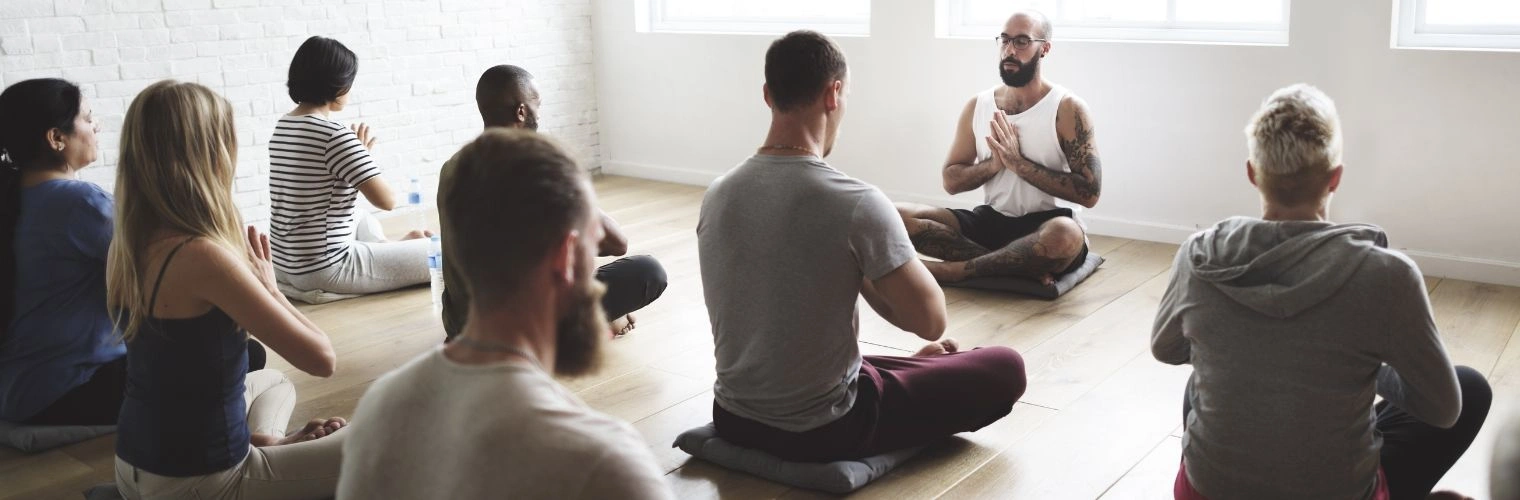 Image of men and women in a yoga class with their legs crossed in the prayer pose, taking direction from yoga instructor up front - 12 Step Recovery Yoga - Ingrained Recovery