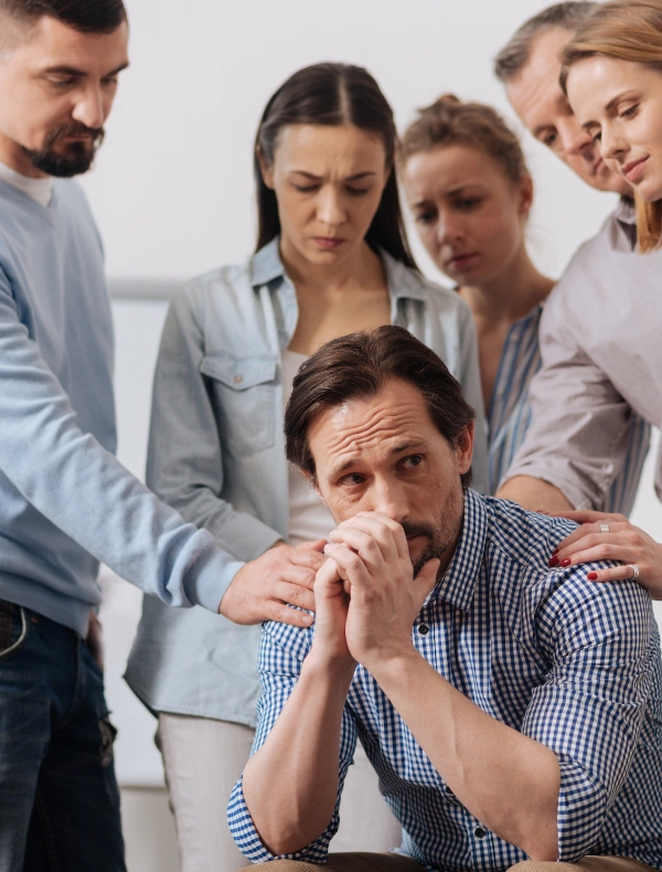 Image of men and women gathered around a somber man at an AA meeting