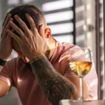 Image of a man sitting at a table in front of a glass of wine as he holds both hands on his head - Drinking in the morning before work - Ingrained