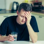 Image of a man sitting at a table with a glass of water in his hand and suffering from a hangover - How Long Does It Take To Sober Up - Ingrained Recovery