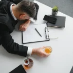 Image of a man passed out on a desk, sitting in front of a legal pad and a glass of whiskey in one hand - Is It Illegal to Drink at Work - Ingrained Recovery