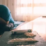 Image of a man sitting with has hands on hi face beside a table with lines of cocaine on it - Why Does Cocaine Make Me Tired - Ingrained Recovery