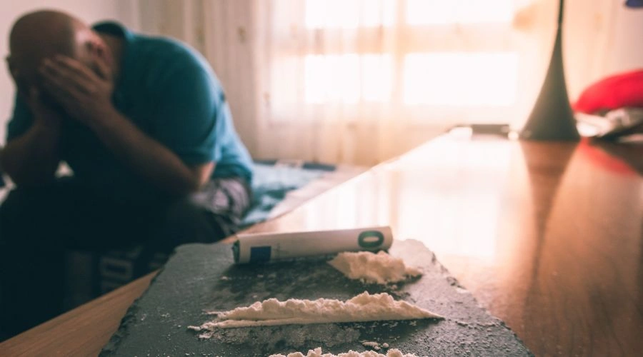 Image of a man sitting with has hands on hi face beside a table with lines of cocaine on it - Why Does Cocaine Make Me Tired - Ingrained Recovery