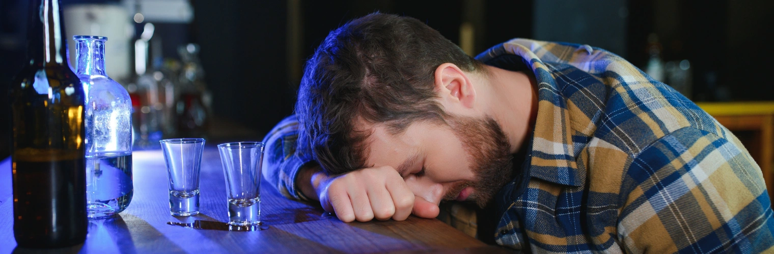 Image of Intoxicated man, passed out with his head down at a table beside bottles of whiskey and empty glasses Image of Intoxicated man, passed out with his head down at a table beside bottles of whiskey and empty glasses