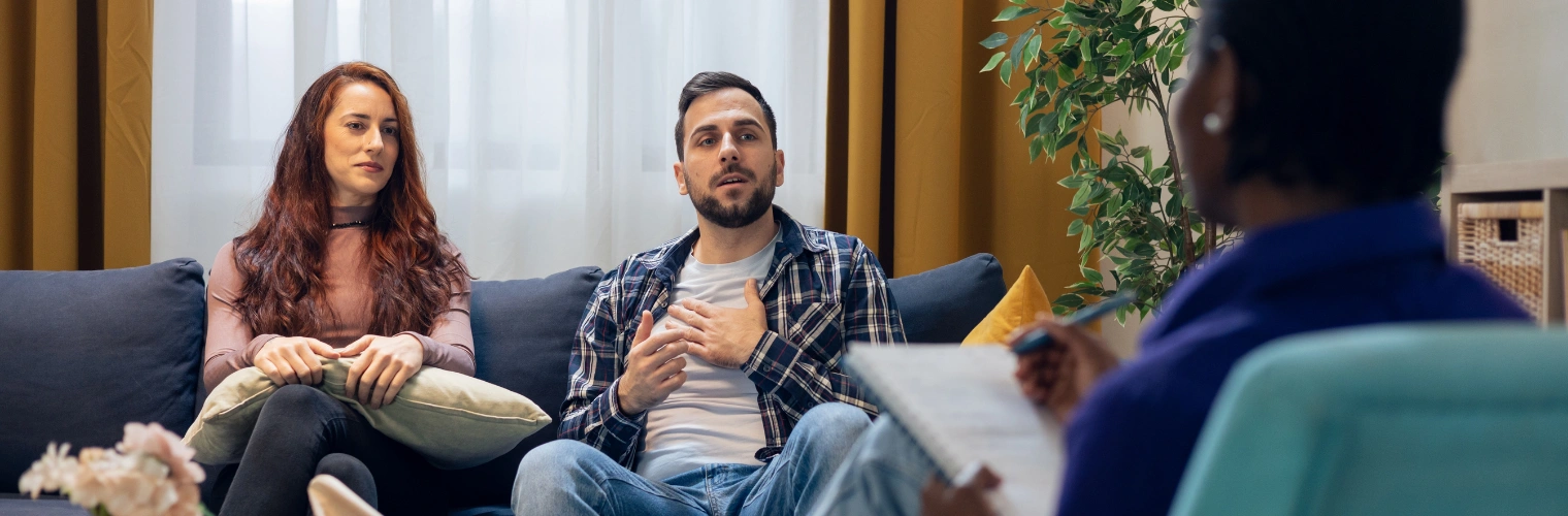 Image of couple engaging in guided therapy to rebuild trust, improve daily problem-solving Image of couple sitting on a sofa and engaging in guided therapy with a counselor to rebuild trust, improve daily problem-solving